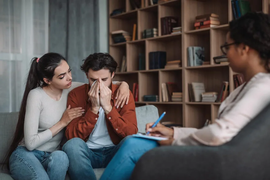 Man struggling with addiction receiving emotional support from partner during counselling session with therapist